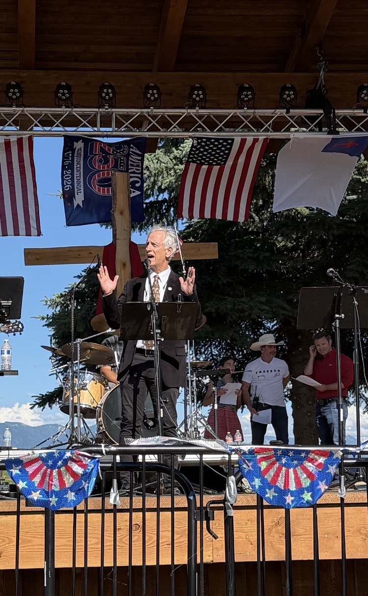 Tracy Sharp is using his hands in the air to emphasize a talking point during his speech at the Faith and Freedom Festival on July 3rd 2026. He is an older man with short white hair and business suit with a red tie.